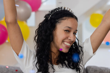 Smiling woman celebrating with balloons, wearing happy birthday headband at home