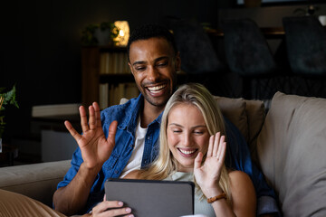Couple on couch video chatting on tablet, smiling and waving at home