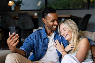 Couple sitting on couch laughing while taking selfie with smartphone at home