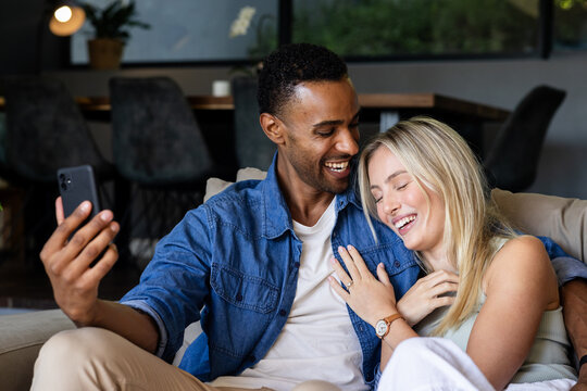 Couple sitting on couch laughing while taking selfie with smartphone at home