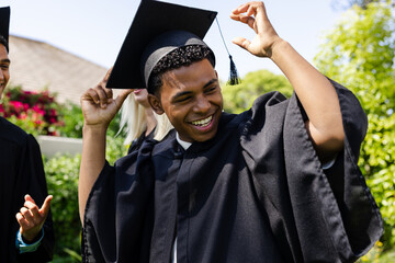 Graduate celebrating outdoors, smiling and adjusting cap with friends nearby