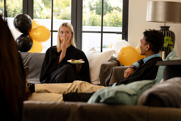 Graduates in gowns relaxing on sofa, enjoying snacks at home celebration