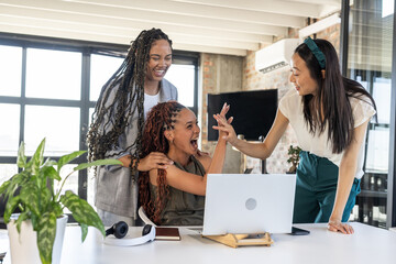 Celebrating success, diverse businesswomen high-fiving at office desk with laptop