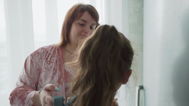 mother in pink robe helps daughter adjust backpack straps by sunny window while boy in grey shirt loads books into backpack ready for school during warm bright kitchen morning routine clip