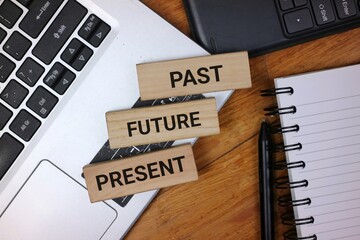 Wooden blocks labeled with 'Past,' 'Present,' 'Future,' placed alongside a laptop and notebook,...