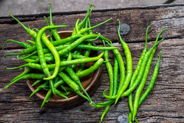 Fresh organic green chili peppers placed in a wooden bowl with some scattered around on rustic wooden surface.