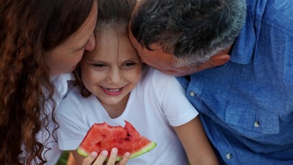 Family enjoys picnic in park. Girl holds juicy slice of watermelon, while mother and father kiss daughter and laugh happily. Concept of family relationships, summer outdoor recreation - Powered by Adobe