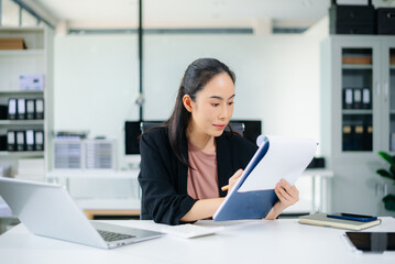 Obraz premium Smiling Asian businesswoman texting on phone at modern office desk with laptop, coffee, and documents