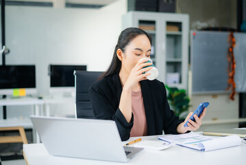 Fototapeta premium Confident beautiful Asian businesswoman typing laptop computer and digital tablet while holding coffee at office