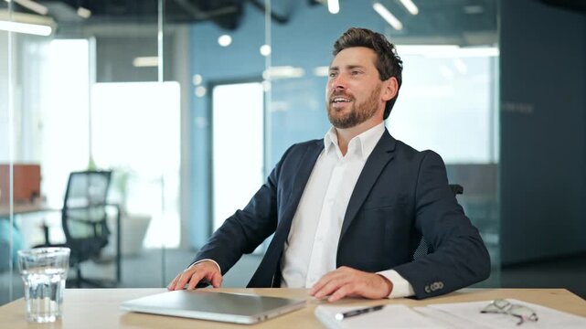 Successful businessman, dressed in a sharp suit, finishes his tasks in a modern office, then leans back feeling content and satisfied with his work.