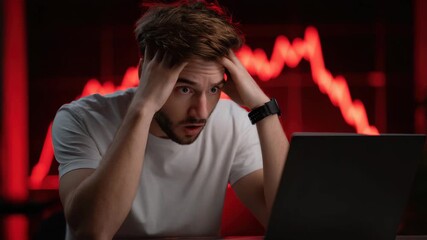 Young man in white shirt stressed and shocked using laptop with red declining stock chart and financial loss, technology and business concepts showing anxiety and worry expressions