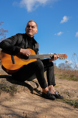 Naklejka premium Beautiful blonde man playing acoustic guitar and singing relaxing enjoying outdoors on sunny autumn day. Artist learning and practicing playing musical instrument. Fall leaves autumnal weather 
