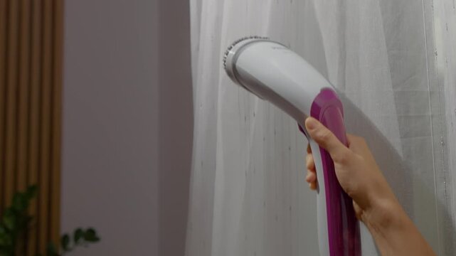 A hand-held steam iron is used to smooth out white drapery, removing wrinkles and creating a crisp, clean look in a comfortable home environment.