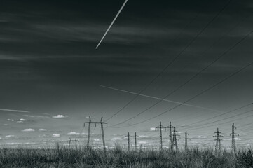 Black and white landscape. Above the wires of a high-voltage power line, the sky is filled with white trails from high-flying airplanes