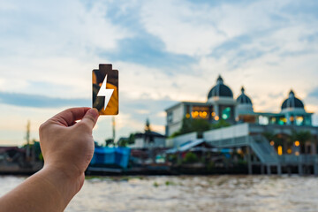 Fototapeta premium Hand holding golden battery icon with lightning bolt against cityscape background, symbolizing energy, electricity, and charging. Perfect for sustainable power, smart city, or eco technology concept.