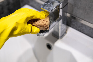 Cleaning the bathroom sink, she is polishing the faucet using a sponge, protective yellow gloves and spray detergent.