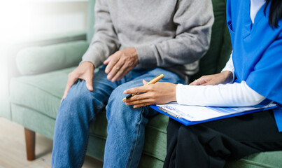 Senior man describes leg pain to nurse at home. Caregiver takes notes during health assessment. Real people in home care, injury checkup, and medical consultation concept.