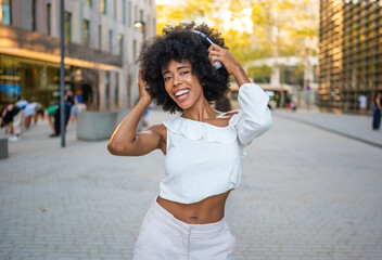 Beautiful young black woman listening to music with headphones and dancing in the city