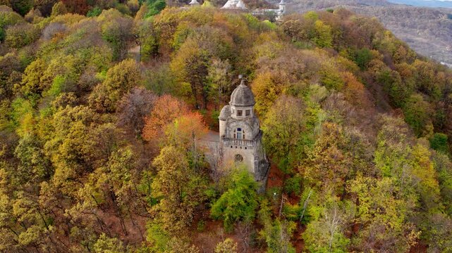 A cinematic aerial shot slowly orbits the Heroes' Monument in Targu Ocna, Romania. The stone tower stands on a hilltop surrounded by a forest of vibrant autumn colors.