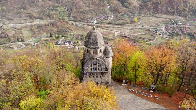 A cinematic aerial shot pulls back to reveal the Heroes' Monument in Targu Ocna, Romania, standing on a hilltop above a forest of vibrant autumn colors and the town below.