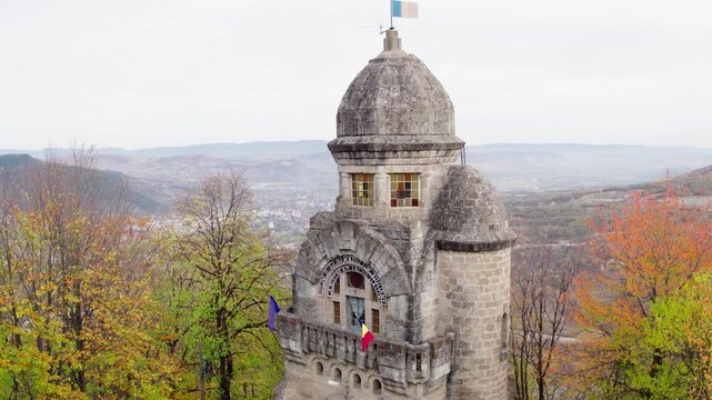 A cinematic close-up aerial shot slowly orbits the Heroes' Monument in Targu Ocna, Romania, showcasing its stone architecture against a backdrop of colorful autumn trees.