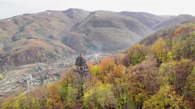 A cinematic aerial shot pulls back to reveal the Heroes' Monument in Targu Ocna, Romania, standing on a hilltop above a forest of vibrant autumn colors and the town below.