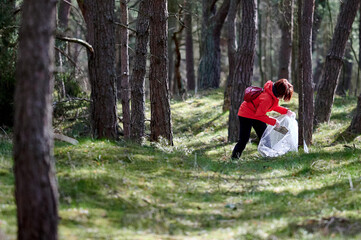 A person in a bright red jacket is seen collecting trash in a forested area. The individual bends over, picking up litter, showcasing an effort in environmental cleanup and nature preservation.