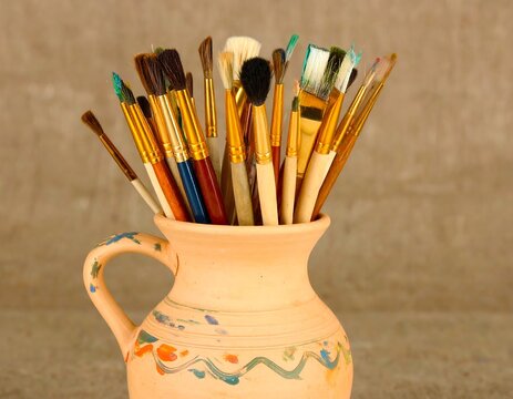 A terracotta pitcher filled with various paintbrushes in varying colors and sizes - Powered by Adobe