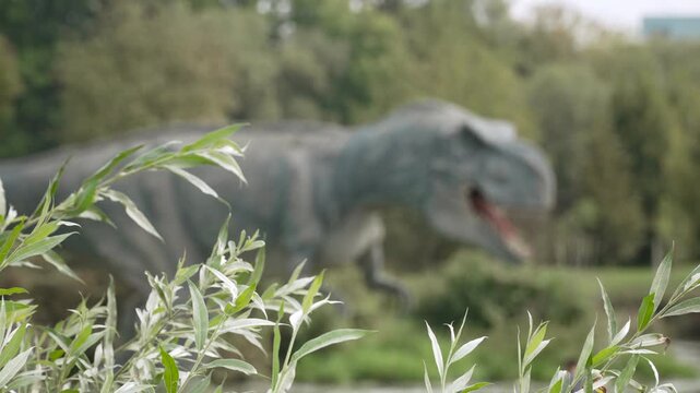 Branches of bushes sway in strong wind as blurred dinosaur model moves among trees in background. Video from amusement park with mechanical animal puppets.