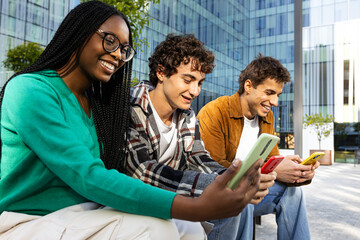 Happy multiethnic students using smartphones sitting on bench in university campus