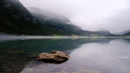 A calm morning at Bergheimsvatnet showcases serene waters reflecting misty mountains. Natural beauty envelops the area, with subtle hues enhancing the tranquil atmosphere. Peaceful vibes abound.