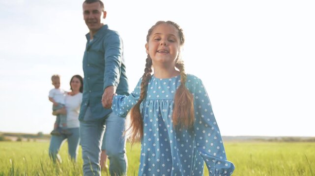 Father leads daughter across field while mother holds son behind in sunny meadow with tall grass and smiling face, family enjoying summer sunlight and playful walk together laughing together happily