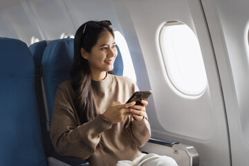 Fototapeta premium An Asian woman sits comfortably on an airplane, holding her smartphone smiling while looking out the window, representing modern travel,connectivity, leisure,the joy of experiencing a flight journey