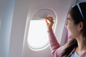 An Asian woman sits near the airplane window, smiling warmly while wearing a casual pink sweater...