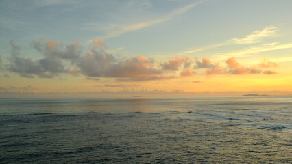A peaceful scene of the calm sea stretching to the horizon under a sky with soft, pastel-colored clouds. A small island is visible in the distance. The image conveys a sense of serenity