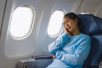 Fototapeta premium An elderly Asian woman sits inside an airplane, suffering from dizziness, nausea, sweating, paleness, headache, fatigue, and drowsiness, experiencing worsen motion sickness during turbulence