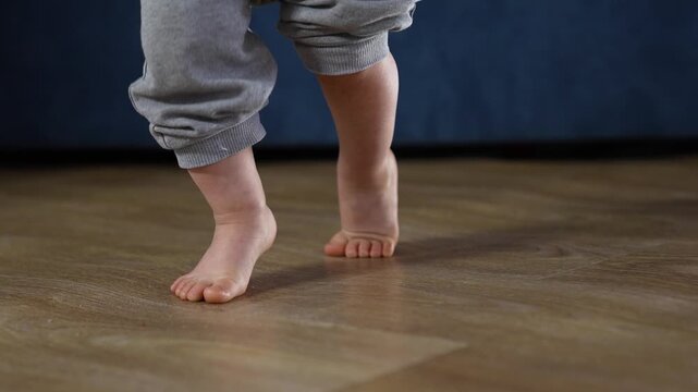 Toddler walking barefoot across wooden floor baby step balance shown as child lifts toe and sets heel with careful step small foot contact on smooth floor in home living area showing steady balance