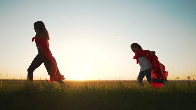 Girls run across grassy field wearing red cape and playful superhero pose at golden sunset, child and girl silhouette on horizon, two children run and play through tall grass under warm sky laughing