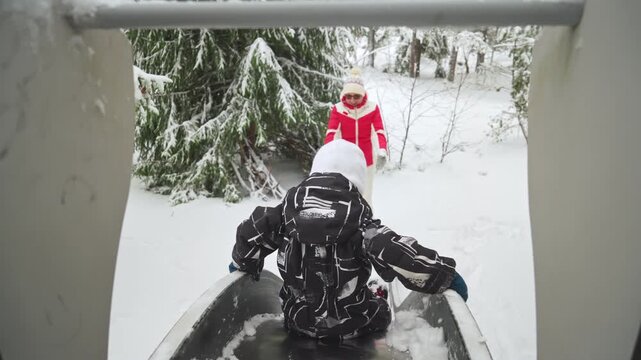 Grandmother catching child on snowy playground slide in Finland. Fun family lifestyle, outdoor winter play, warm clothing, love, joy, and happy childhood emotions.