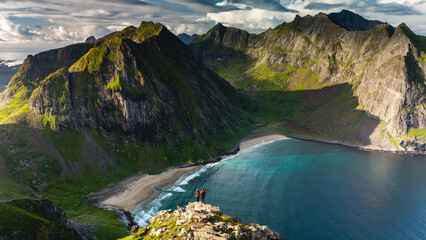 Captivating panorama from the Ryten mountain trail showcases Kvalvika Beachs pristine shoreline, embraced by majestic mountains under a clear sky. Ideal for hiking enthusiasts and nature lovers.