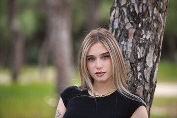 Portrait of beautiful young blonde woman in black shirt leaning against a tree trunk in the forest. The woman is serious and staring.