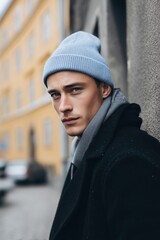 A young man wearing a light blue beanie mockup and a dark coat, standing against a textured wall, with a serious expression, urban background