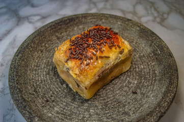 Golden layered pastry slice with flaky crust and creamy filling, topped with chocolate sprinkles on dark plate ideal for highlighting baked texture, sweet detail, and dessert appeal.