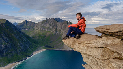 A hiker rests on the edge of a rock promontory, enjoying the stunning panorama of Lofotens mountains, beaches, and crystal-clear waters during a tranquil moment in Norways nature.