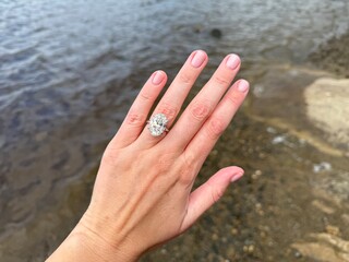 A landscape photo showing a woman hand with an oval diamond engagement platinum ring, resting near water with soft natural daylight glow.