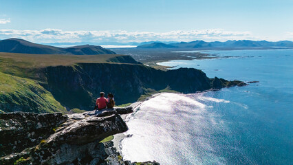 Two hikers enjoy stunning coastal views from the summit of Matind Mountain. The clear blue sky...