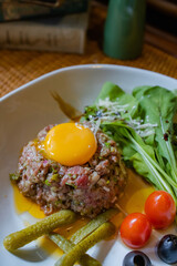 Gourmet Steak Tartare: A meticulously crafted steak tartare, adorned with a vibrant egg yolk, presented alongside fresh greens, juicy cherry tomatoes, savory pickles, and olives.