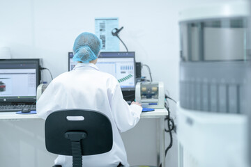 From behind, a lab professional in a hair net works at a computer, analyzing patient test results and monitoring medical equipment in a modern hospital laboratory.