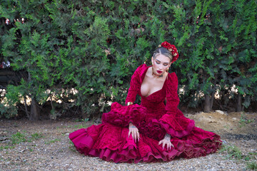 Fototapeta premium Young, beautiful, blonde woman in red flamenco dress, flowers in her hair and gold jewelry, performing flamenco poses sitting on the ground with green plants in the background. Concept dance, art.