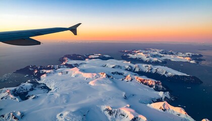 Aerial view of snow-capped Antarctic mountains and islands at sunset, with an airplane wing in the foreground
